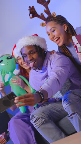 Vertikale-Videostudioaufnahme-Von-Freunden-Der-Generation-Z-Zu-Weihnachten,-Die-Mit-Weihnachtsmütze-Und-Rentiergeweih-Auf-Dem-Sofa-Sitzen-Und-Ein-Selfie-Mit-Dem-Mobiltelefon-Machen-2